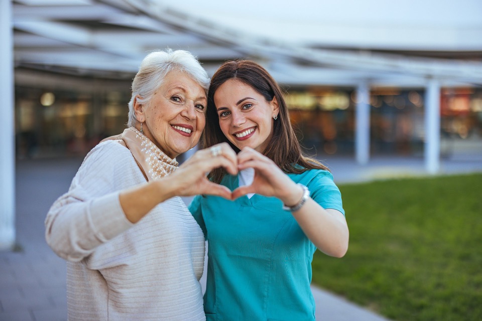 Seniorin und ihre Betreuungsperson formen ein Herz mit den Händen und stehen für Sicherheit, Fürsorge und Unterstützung im Alltag.